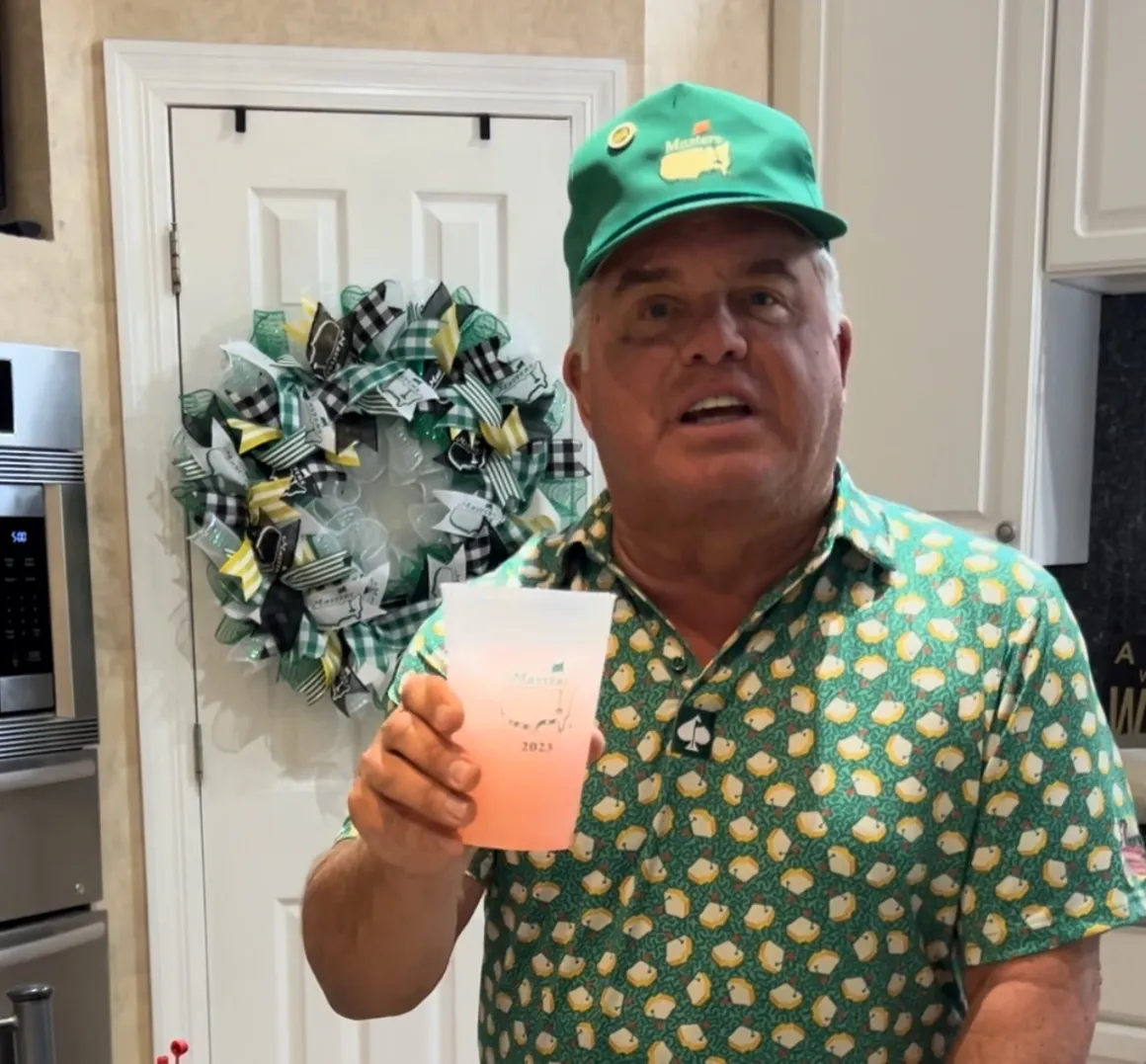 Man holding drink in kitchen, wearing green hat.